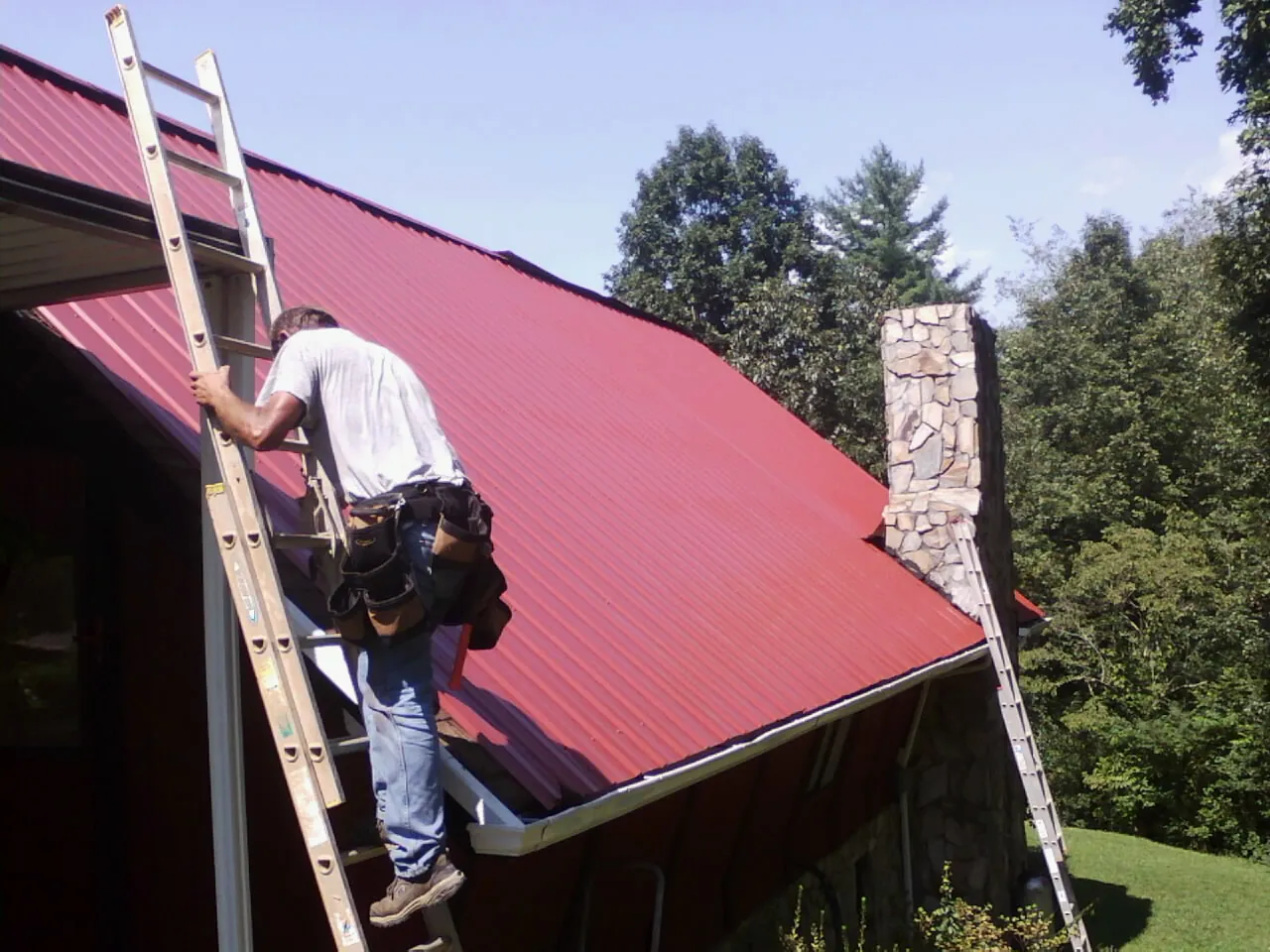 Roofer on a ladder beside a red metal roof and stone chimney — Soffit & Fascia Repair by Roofing Brothers in Longmont